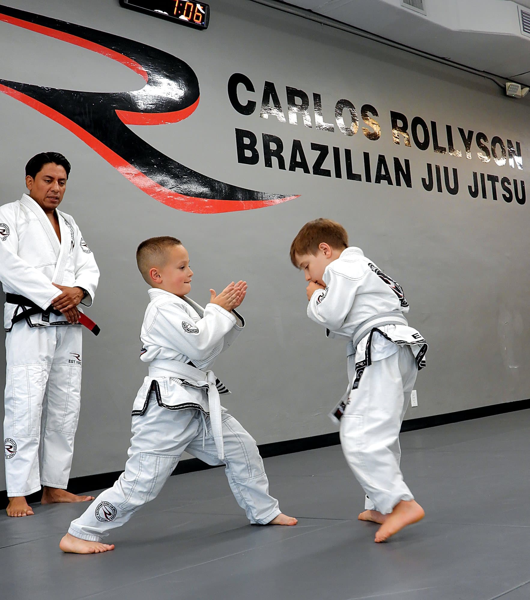 Instructor watching two kids drill a technique in front of the Carlos Rollyson logo wall